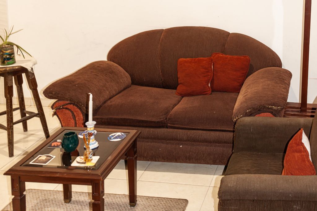 A brown sofa with two red cushions, a wooden coffee table with a vase and a candle, and a small plant on a side table.