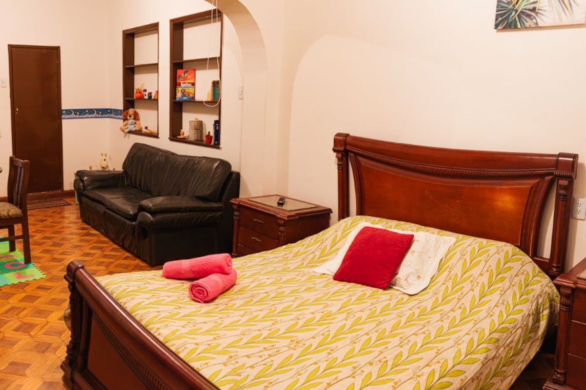 A bedroom with a wooden bed, a leather sofa, a small bedside table, and a wall-mounted shelf with various items.