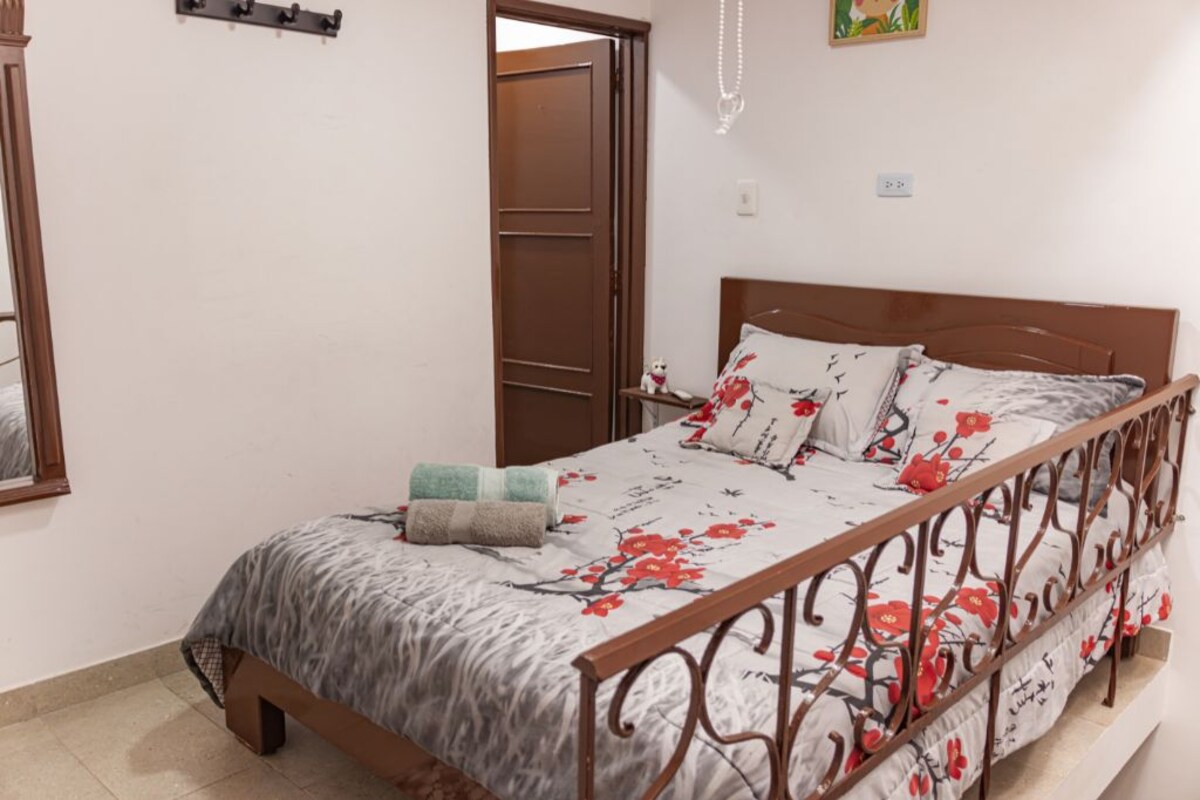 A bedroom with a bed, a wooden door, a wall-mounted light fixture, and a small decorative item on the nightstand.