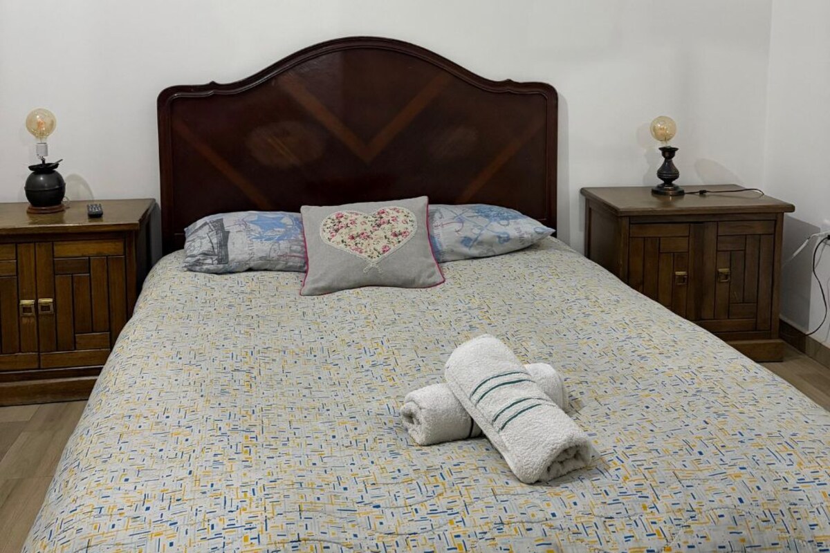 A neatly made bed with a patterned bedspread, a heart-shaped pillow, and rolled towels on the bed.