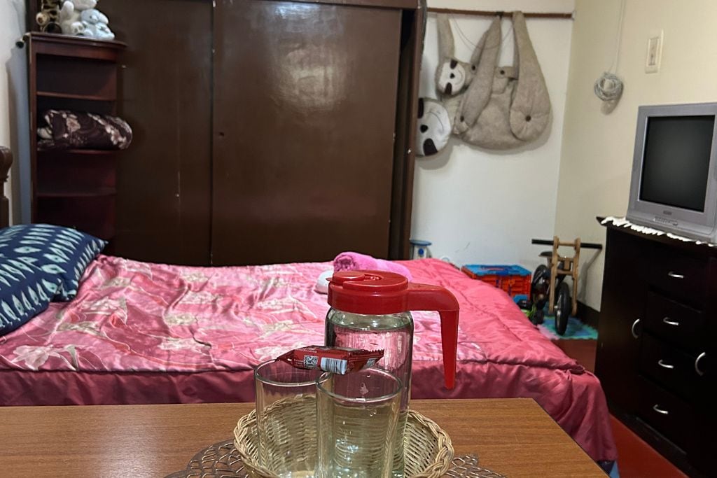A bedroom with a bed, a wooden dresser, a television, and a wall-mounted bag.