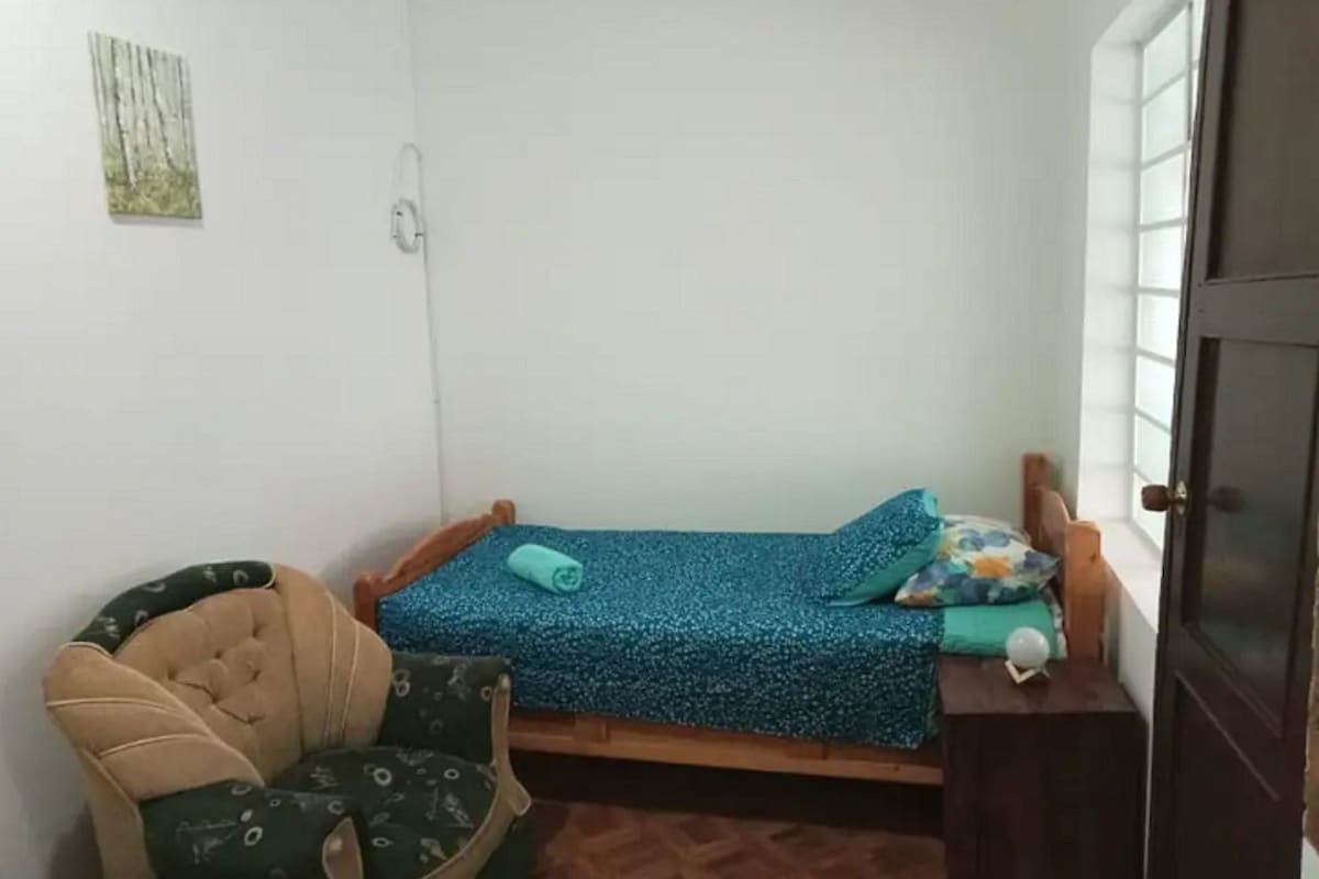 A small, single-bed room with a wooden headboard, a chair with a cushion, a small table, and a window.