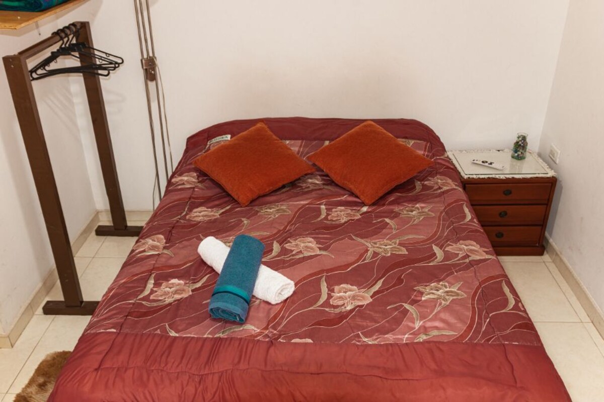 A bed with a floral bedspread, two pillows, a rolled-up towel, and a small bedside table with a remote control.