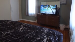 Ein Schlafzimmer mit einem Bett, einem Fernseher auf einer Kommode, einem Fenster mit Vorhängen und einem Gemälde an der Wand.