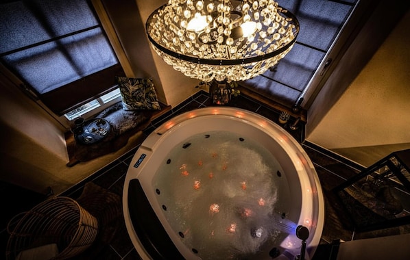 Un bain à remous surmonté d’un lustre, une fenêtre avec des stores et une petite table agrémentée d’un objet décoratif.