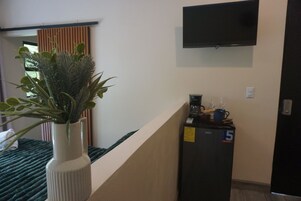 Habitación de hotel con una cama, una planta en un jarrón blanco, un televisor de pantalla plana montado en la pared y un minibar con cafetera y vasos.
