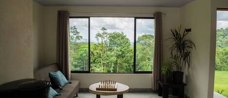 Una habitación con un ventanal grande, un sofá, una mesa con piezas de ajedrez y plantas en macetas.