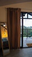 Una habitación con un espejo grande, una cama con ropa de cama azul, un ventilador de techo y vistas a un balcón con una silla y follaje exterior.