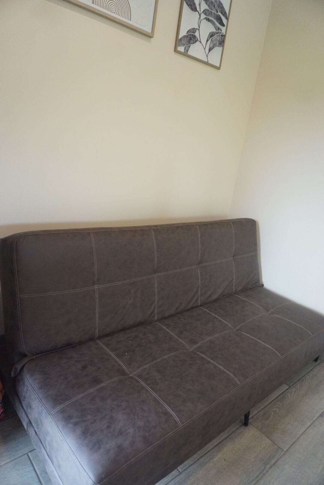 A dark brown sofa with a cushioned backrest and armrests, positioned against a plain wall with two framed artworks above it.