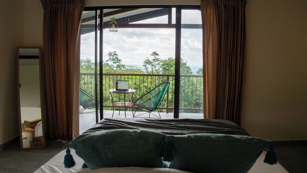 Un dormitorio con una ventana grande, una cama, una silla y un ordenador portátil sobre una mesita.