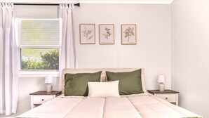 Un dormitorio con cama, dos mesitas de noche, una ventana con cortinas y tres láminas enmarcadas con motivos botánicos en la pared.