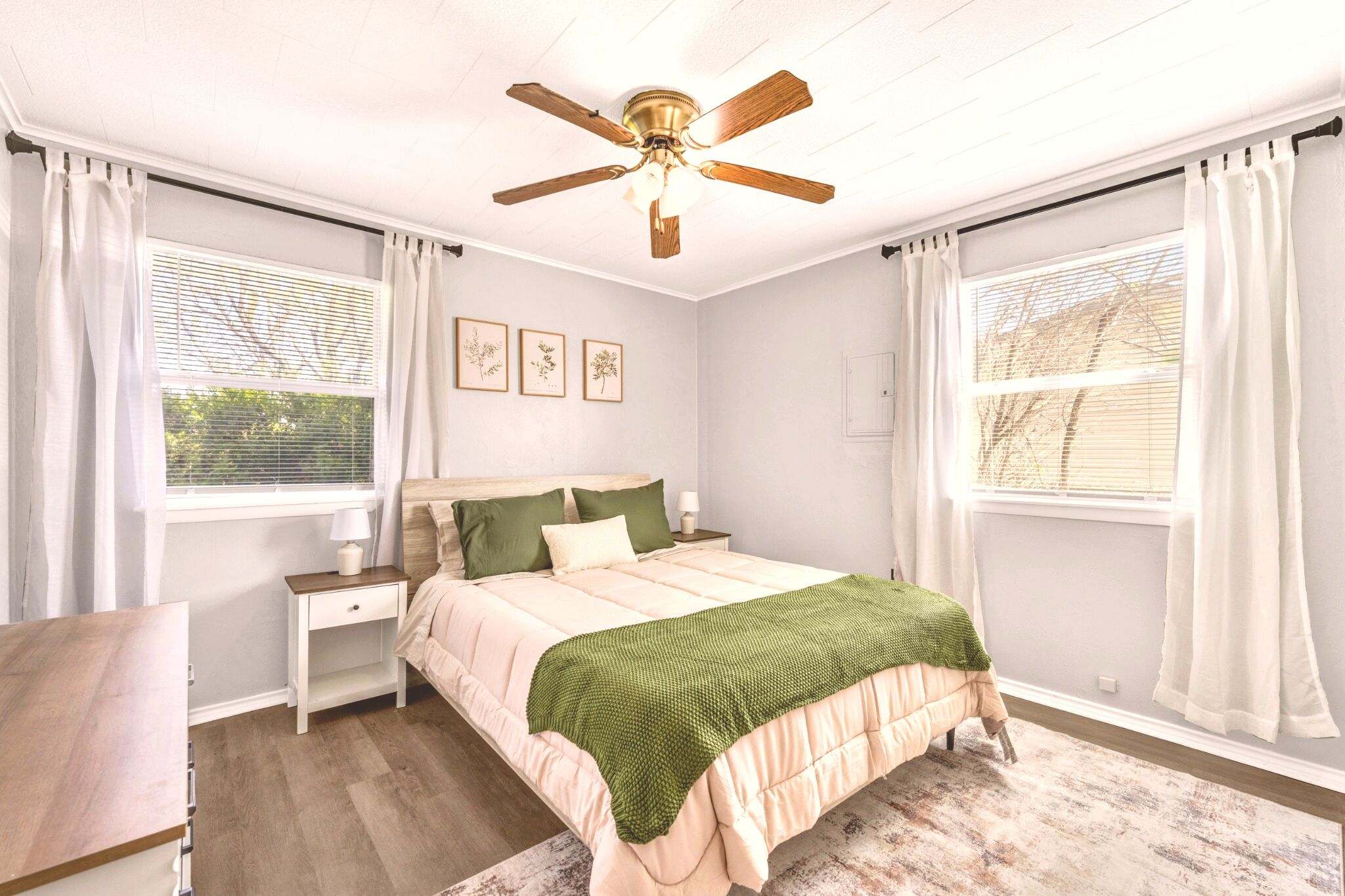 A bedroom with a bed, nightstand, ceiling fan, and windows with curtains.