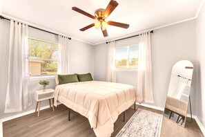Une chambre à coucher comprenant un lit, une table de chevet, un ventilateur de plafond, une fenêtre avec des rideaux et un miroir.