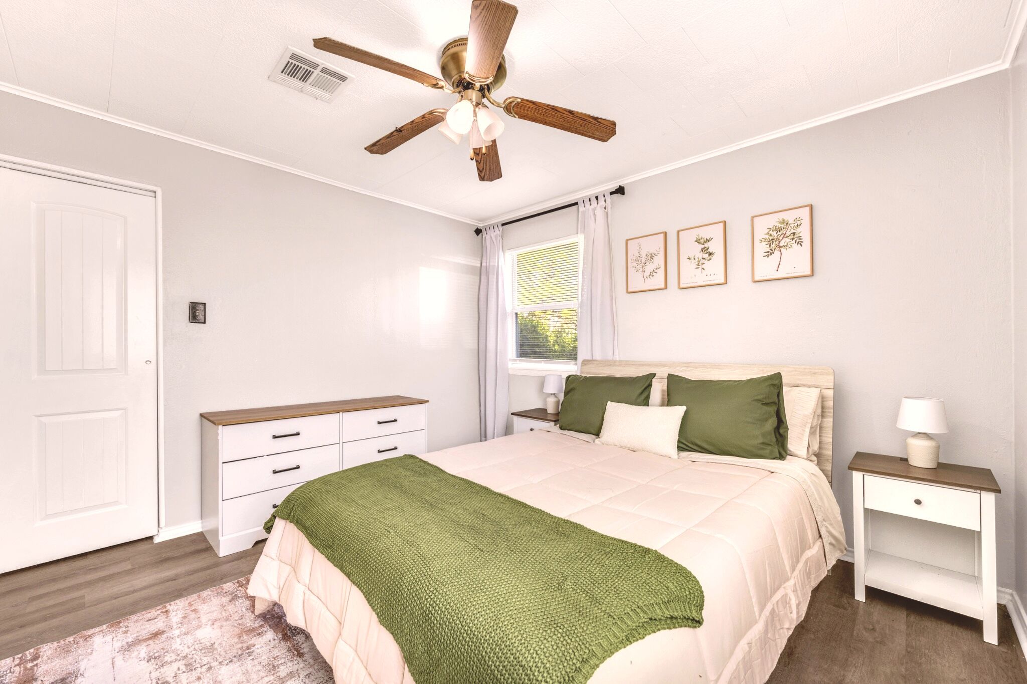 A bedroom with a bed, nightstand, dresser, ceiling fan, window with curtains, and framed botanical prints on the wall.
