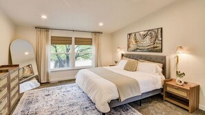 Ein Schlafzimmer mit einem großen Bett, einem Fenster mit Vorhängen, einem Nachttisch mit Lampe und einem an der Wand hängenden Kunstwerk.