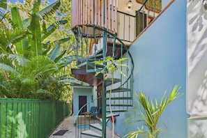 Un escalier en colimaçon menant à un balcon, entouré d’une végétation luxuriante et bordé d’un mur bleu.