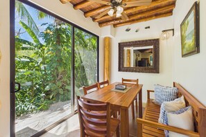 A wooden dining table with chairs, a large mirror, and a view of lush greenery outside.