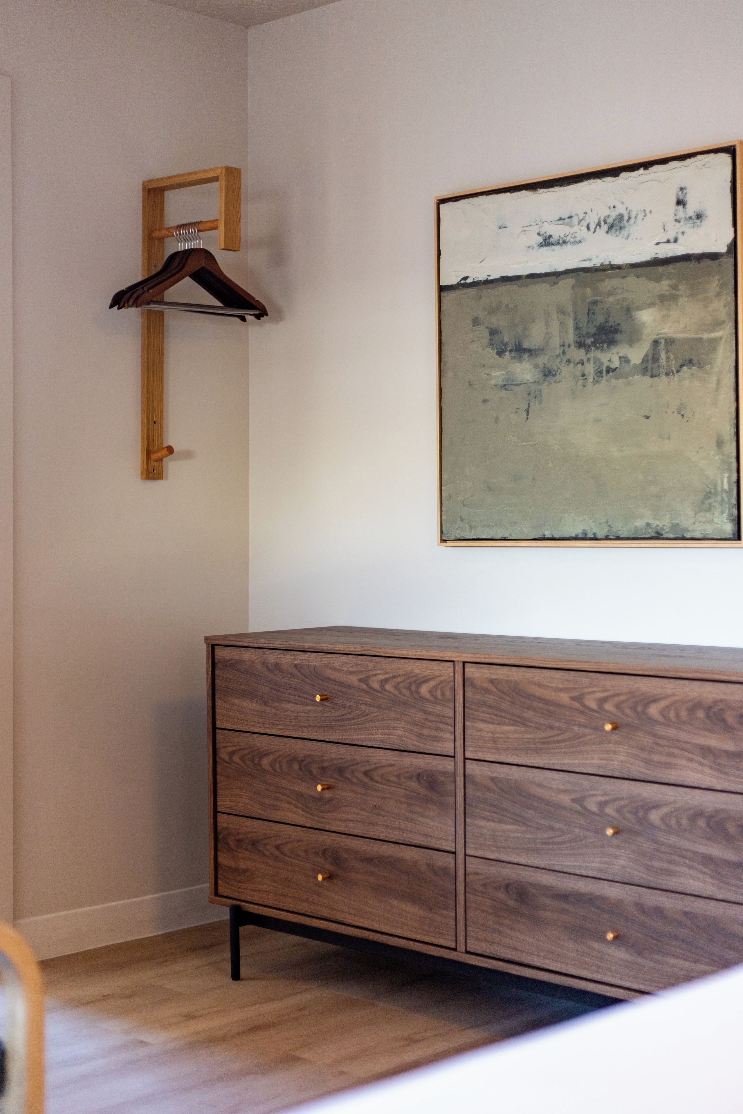 A wooden dresser with drawers, a wooden coat rack, and a large abstract painting on the wall.