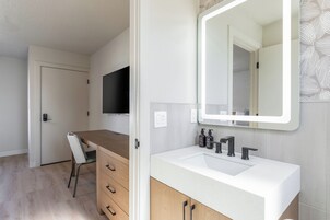 Un baño moderno con un lavamanos blanco, grifo negro y un espejo grande. Se observa un mueble de madera con cajones y una silla en la esquina.