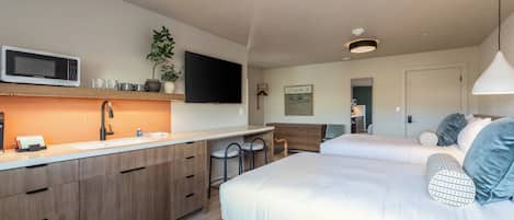 Una habitación de hotel moderna con kitchenette, un televisor de pantalla plana, una cama con almohadas y una mesita de noche.