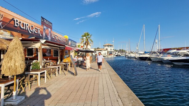 Eine Marina mit anlegenden Booten, ein Restaurant namens 'Burger Restaurant' auf der linken Seite und ein klarer, blauer Himmel.