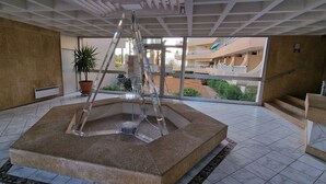 Ein moderner Innenbereich mit einem zentralen Wasserspiel, einem Blumentopf und Blick auf ein mehrstöckiges Gebäude mit Balkonen.
