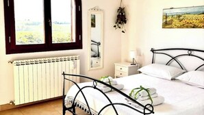Een slaapkamer met een bed, een raam, een radiator, een spiegel, een nachtkastje en een schilderij aan de muur.