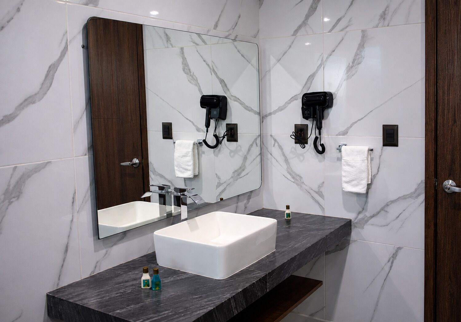 A modern bathroom with two sinks, a large mirror, and a hairdryer mounted on the wall.