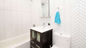 A modern bathroom with a white herringbone-patterned wall, a black cabinet with a white countertop, a white toilet, and a blue towel hanging on the wall. - Luxury 2BR/1BA - 10 min to NYC (Hoboken)