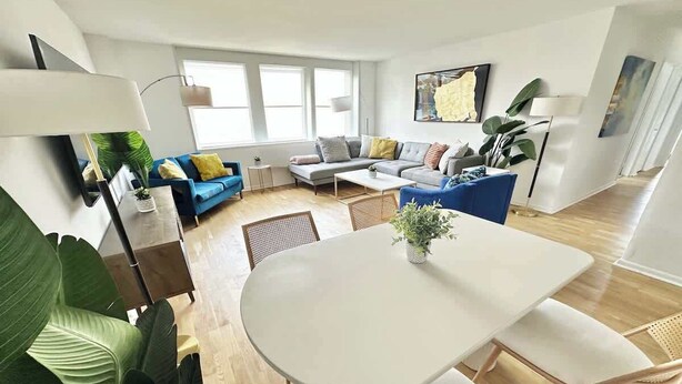A modern living room with a white dining table, blue sofas, and large green plants. - Luxury 2BR/1BA - 10 min to NYC (Hoboken)