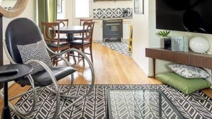 Una sala moderna con un sofá de cuero negro, un comedor con mesa y sillas de madera, un televisor negro sobre un mueble de madera, y baldosas de suelo estampadas.