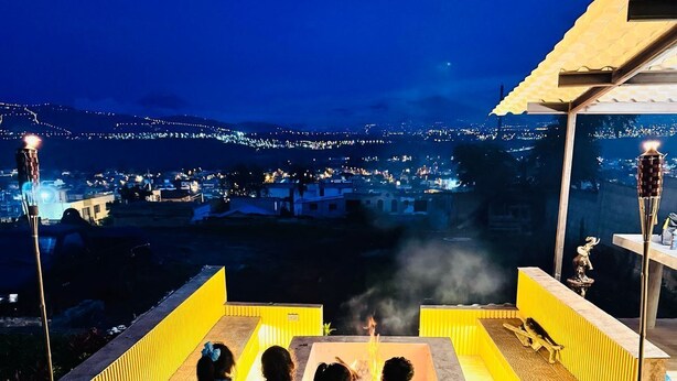 Une scène nocturne avec un espace aménagé autour d’un foyer, une vue sur un paysage urbain et un premier plan verdoyant.