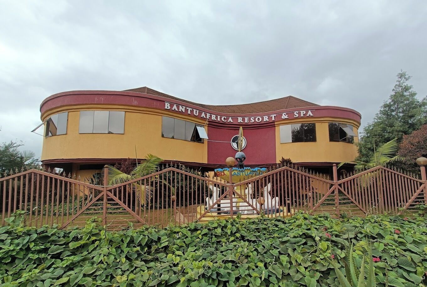 En byggnad med skylten ”Bantuafrica Resort & Spa”, en böjd fasad, en central entré med trappsteg och ett dekorativt staket framför.