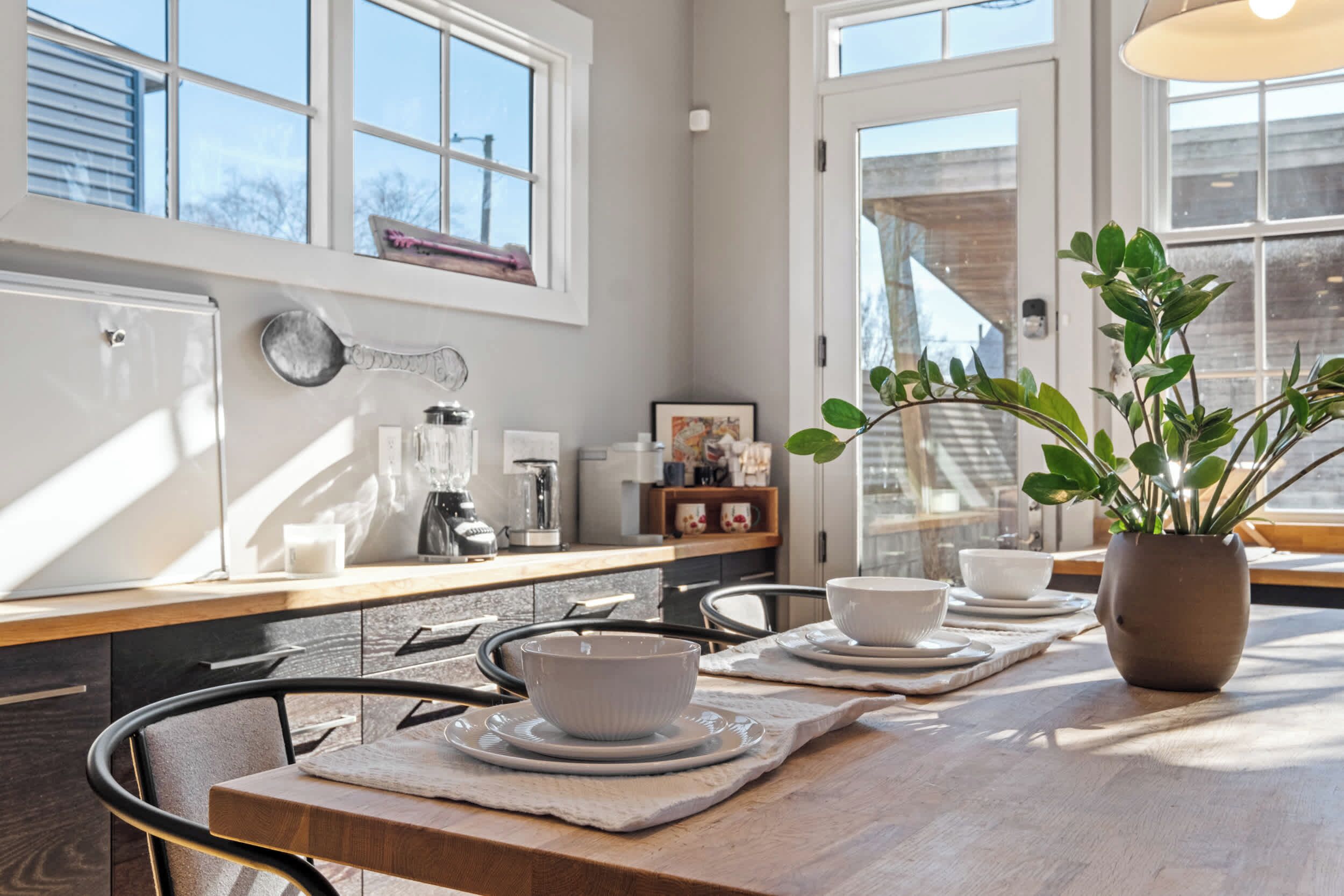 Une cuisine avec une table en bois dressée pour quatre personnes, une plante en pot et une fenêtre donnant sur l’extérieur.