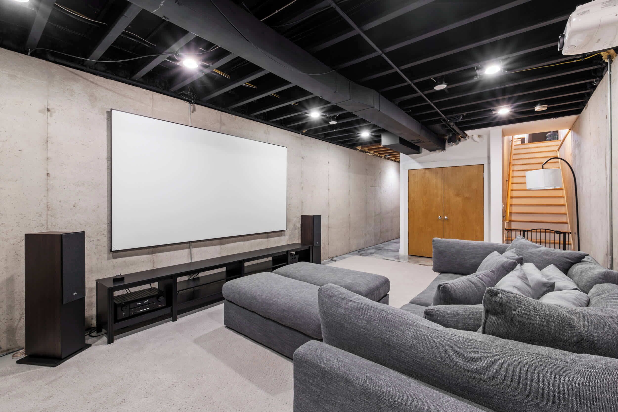 Una sala home theater con un grande schermo bianco, un mobile in legno scuro e sedute rivestite in grigio.