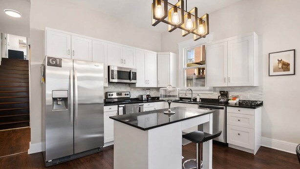 Una cocina moderna con armarios blancos, electrodomésticos de acero inoxidable, una isla central con taburetes y una lámpara colgante.
