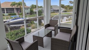 Un balcone con mobili in vimini, un tavolino in vetro e vista su una piscina e delle case.