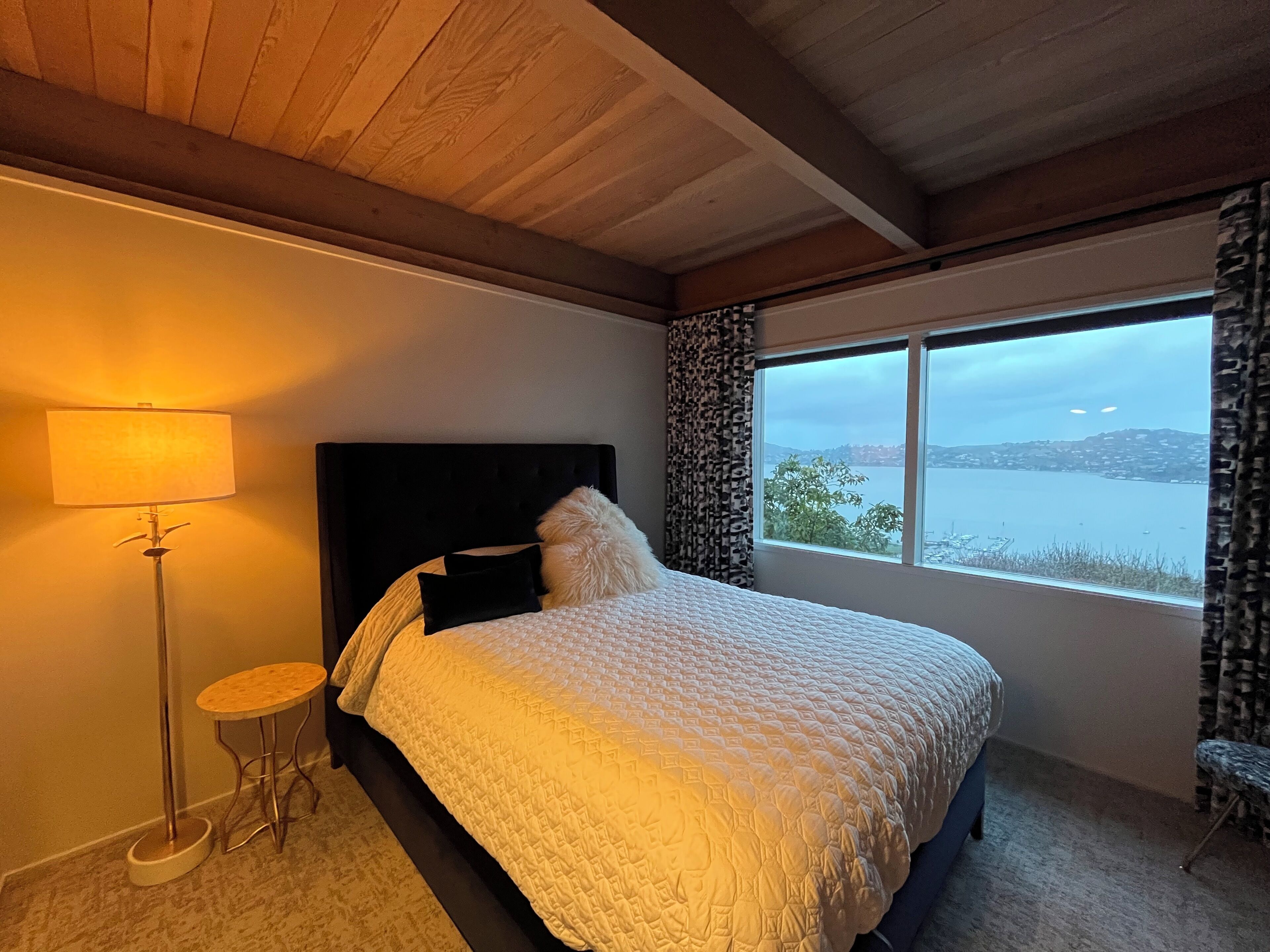 A bedroom with a large bed, a nightstand, a lamp, and a view of a body of water through a window.