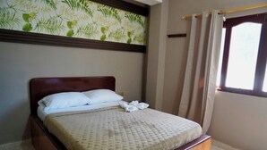 A neatly made bed with a patterned bedspread and pillows, a wooden headboard, a window with curtains, and a wall with a leaf-patterned wallpaper. - Hotel Lua (Santa Marta)