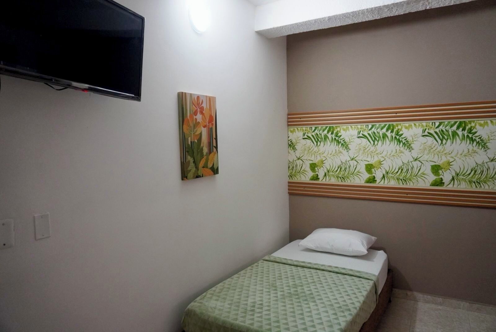 Una habitación pequeña y sencilla con una cama, un televisor de pared y un cuadro con motivos florales.