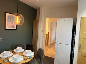 Un comedor con una mesa preparada para cuatro personas, un refrigerador y un pasillo que lleva a una escalera.