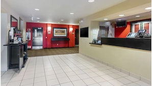 Un espace d’accueil avec un mur rouge, un panneau d’affichage indiquant « Bienvenue », un comptoir d’accueil, une machine à café et un écran de télévision.