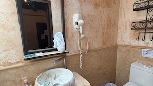 Un baño con lavamanos de mármol, un espejo, un secador de pelo, un inodoro y un estante en la pared.