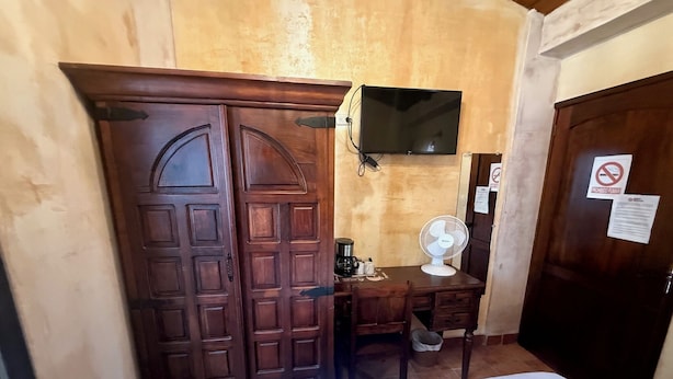 Una habitación con puerta de madera, un escritorio con ventilador, televisor en la pared y una cama.