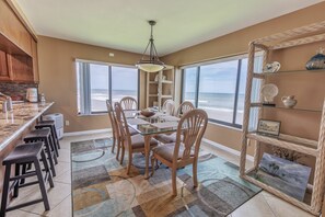 A dining area with a view of the beach, a kitchen with a marble countertop, and wicker shelves. - Oceanview Condo on No-Drive Beach | Balcony, Pool, Peaceful Escape (New Smyrna Beach)