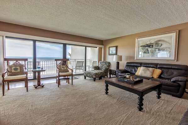 A spacious living room with a large window, a black leather sofa, a wooden chair, a patterned armchair, and a small round table. - Oceanview Condo on No-Drive Beach | Balcony, Pool, Peaceful Escape (New Smyrna Beach)