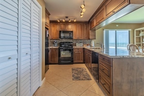 A kitchen with wooden cabinets, a microwave, stove, and a large window overlooking the ocean. - Oceanview Condo on No-Drive Beach | Balcony, Pool, Peaceful Escape (New Smyrna Beach)