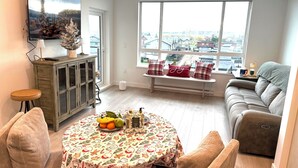 Un salon avec une grande fenêtre, une table ronde recouverte d’une nappe festive, un canapé et un meuble.