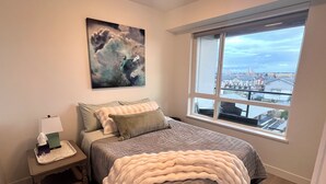 Une chambre avec une grande fenêtre, un lit recouvert d’une courtepointe, une table de chevet avec une lampe et un tableau accroché au mur.
