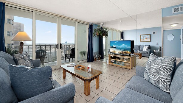Uma sala de estar moderna com uma televisão grande, assentos confortáveis, uma varanda com vista e uma mesa de centro com tema praiano.
